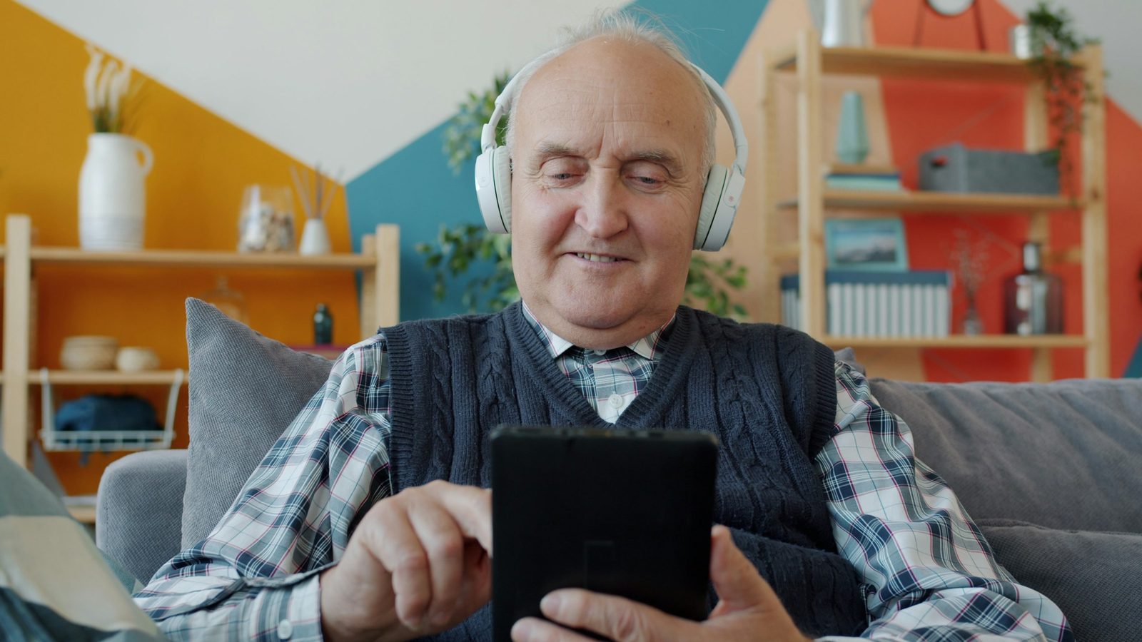 Ein älterer Mann mit Kopfhörern sitzt zu Hause auf dem Sofa, nutzt ein Tablet und lächelt.